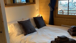 A bedroom with a wooden headboard, a large window with a view of a snowy landscape, a bed with white linens and dark pillows, and a small bedside table with a lamp.