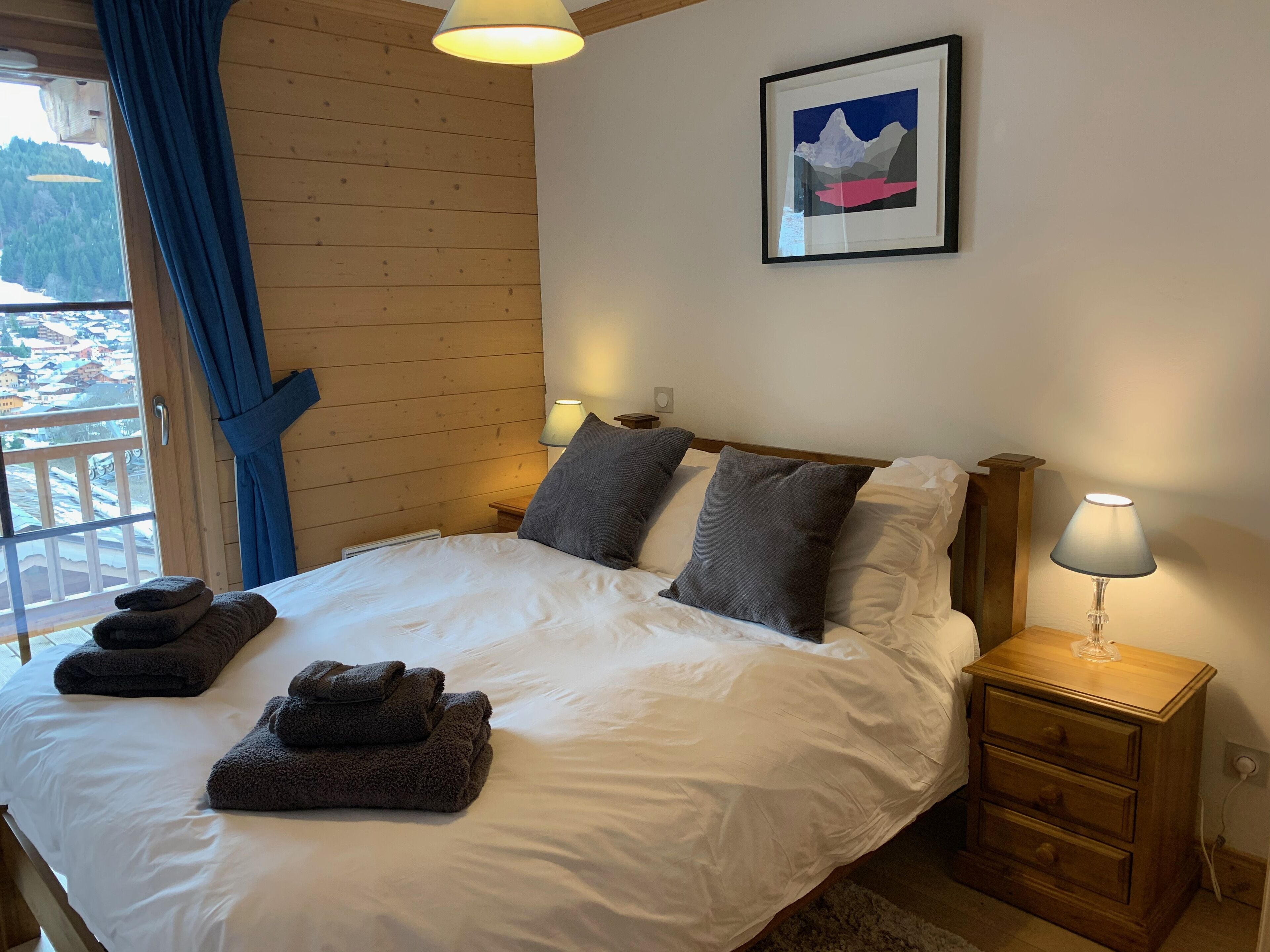 A neatly made bed with white linens and two grey pillows, a wooden nightstand with a lamp, and a framed picture on the wall.