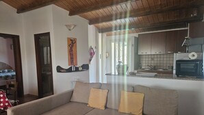 A living room with a grey sofa, wooden ceiling beams, a kitchen with a microwave, and a window with curtains. - Villa surrounded by greenery with private pool  (Palazzolo Acreide)