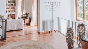 A spacious room with high ceilings, exposed wooden beams, a large window, a white wall with a decorative metal object, and a view of a hallway with a bed and a radiator.