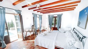 A bedroom with a large bed, wooden beams, a ceiling fan, and a view of the outdoors.