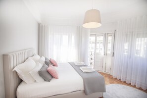 A bedroom with a large bed, white curtains, a hanging light fixture, and a view of the outside.