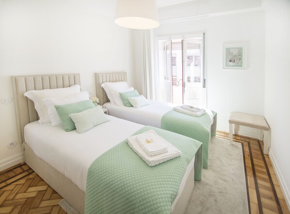 A hotel room with two beds, each with green bedding and white pillows. There is a small bedside table with a lamp, and a framed picture on the wall.