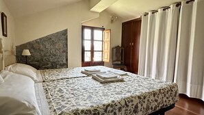 A bedroom with a large bed, a stone wall, a wooden door, and a window with curtains. - Rural Accommodations Casa Fabián, La Tenada (Pet Friendly) (Castrillón)