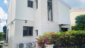 A modern building with white tiled exterior, air conditioning units, and a small garden with pink flowers. - Luxury Villa With Private Pool, Landscaped Garden And Sea Views (Pissouri Bay)
