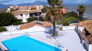 Una piscina con solárium de baldosas, una palmera y vistas a edificios y al mar.