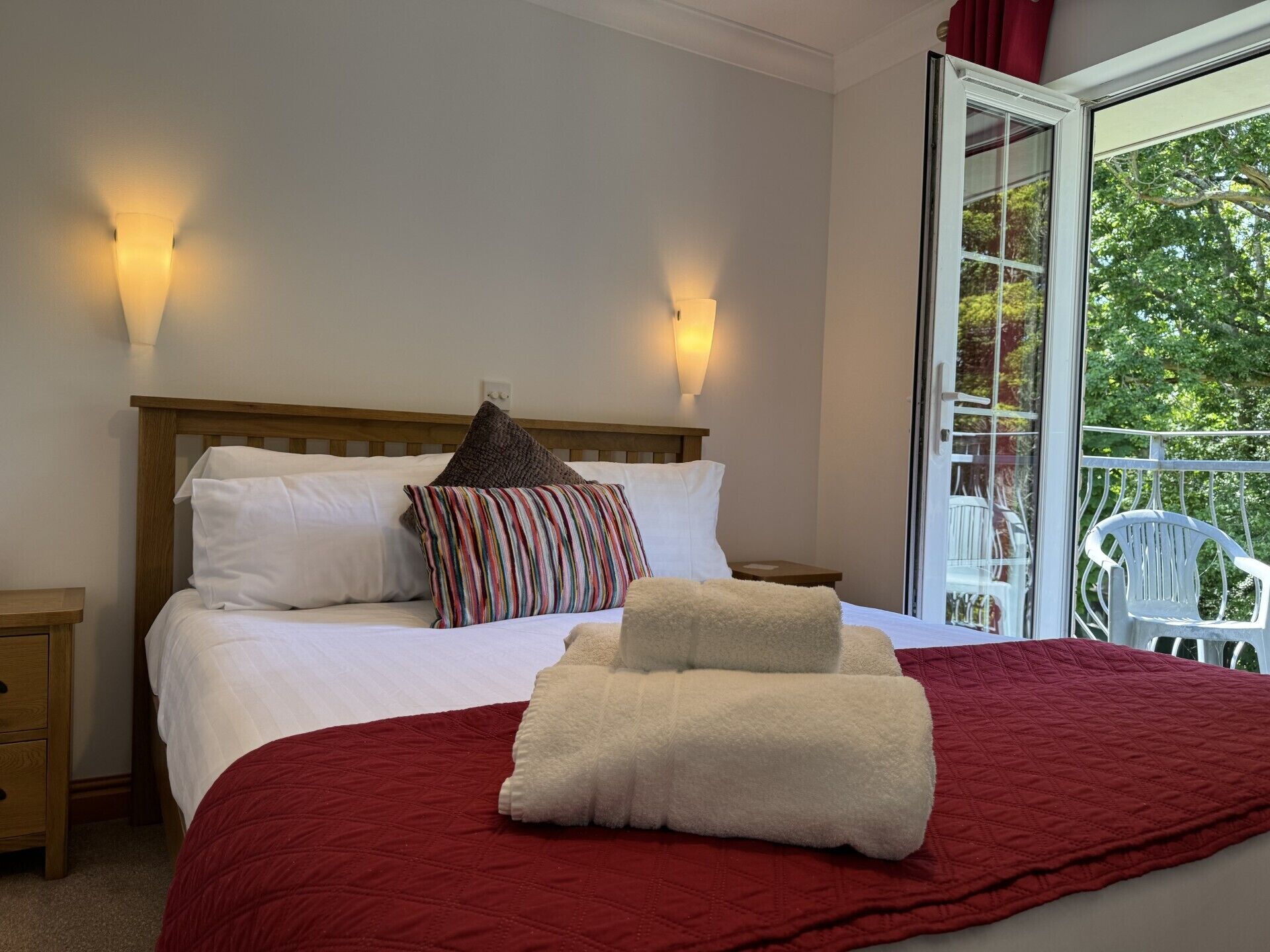 Un dormitorio con cama de madera, ropa de cama blanca, una manta roja y un balcón con sillas blancas.