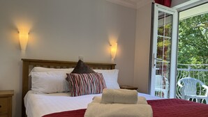 Une chambre avec un lit en bois, du linge de lit blanc, une couverture rouge et un balcon avec des chaises blanches.