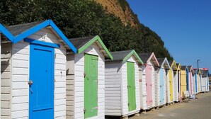 Une rangée de cabanes de plage colorées, dotées de portes, dont certaines sont surmontées de numéros, le long d’une plage de sable, avec une falaise en arrière-plan.