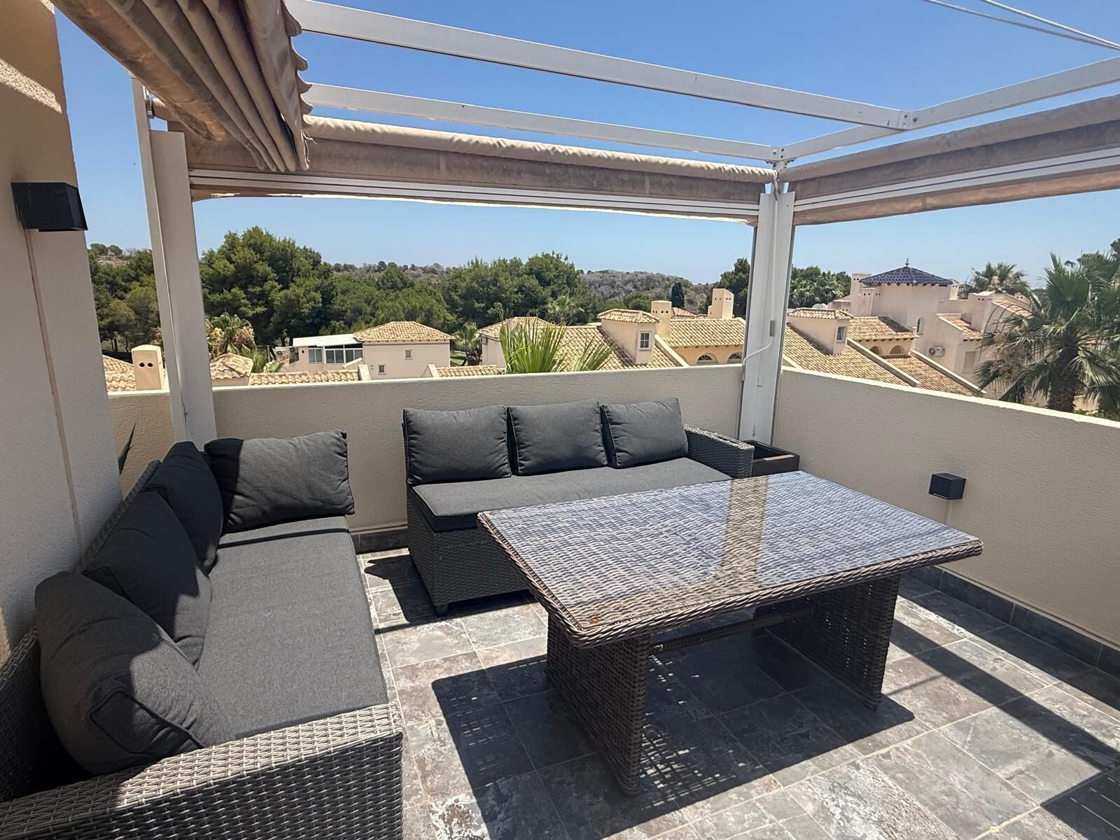 Un área de estar al aire libre con techo, que cuenta con una mesa y sofás, con vista a un barrio residencial.