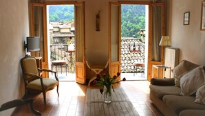 A room with a wooden floor, a sofa, a chair, a coffee table with a vase of flowers, a television on a stand, and a balcony with a view of greenery and buildings. - Historic House With Stunning Mountain Views in the heart of the village      (Pigna)
