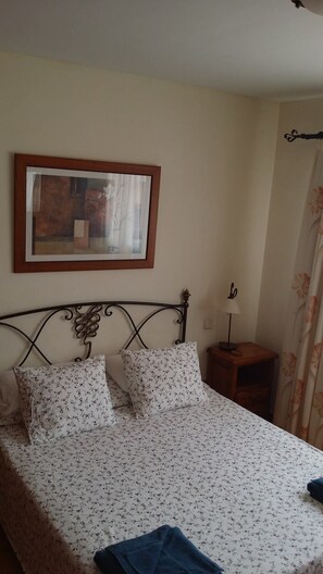 Un lit avec une literie à motifs floraux, un tableau encadré au mur, une lampe de chevet et un rideau.