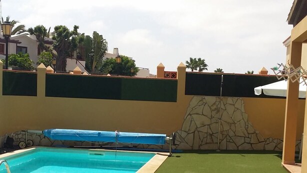 Piscina con tres tumbonas sobre césped artificial, rodeada por un muro de piedra y un edificio de fachada amarilla.