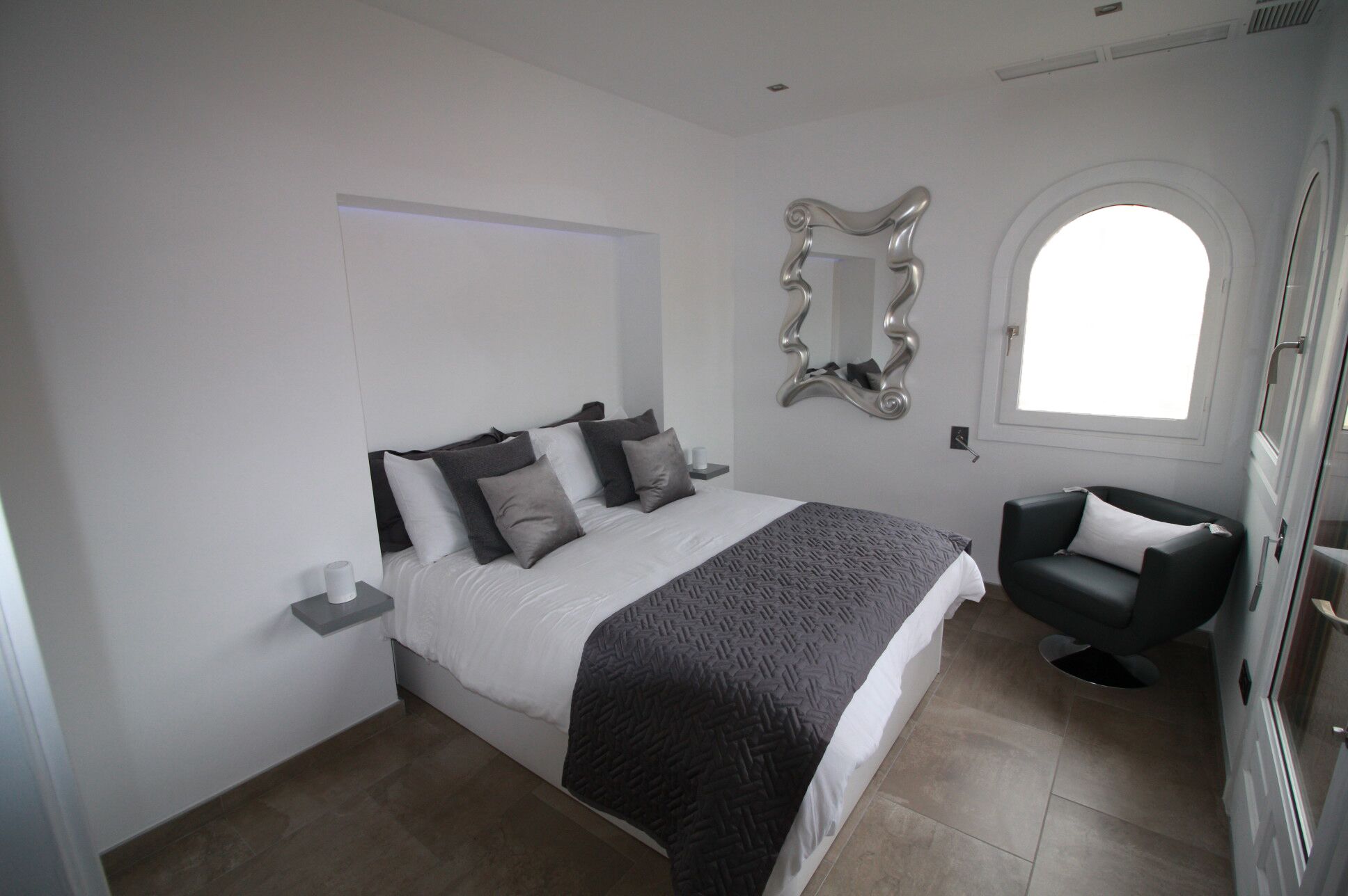 A modern bedroom with a bed, a chair, a mirror, and a window.