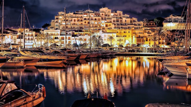 Un port de plaisance avec des bateaux amarrés, un ensemble de bâtiments au bord de l’eau et une étendue d’eau calme qui reflète les lumières.
