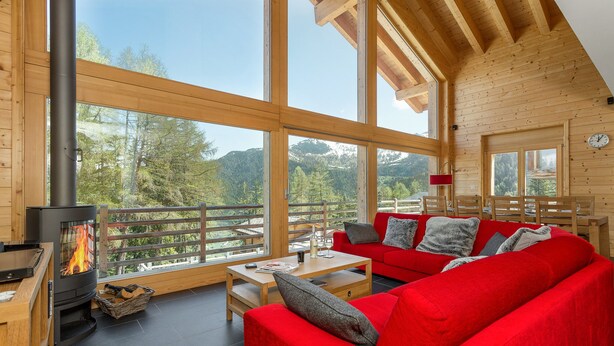 A cozy wooden cabin interior with a red sofa, a fireplace, and large windows offering a view of the forest. - Chalet Anna - La Tzoumaz, 4 Vallees (La Tzoumaz)