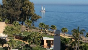 A coastal view with a sailboat on the water, palm trees, and a building with a modern design. - Bahia Beach Front Apartment - Close to Marbella - Free WiFi (Estepona)