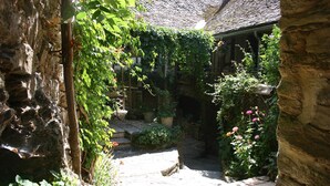 Un sentier de pierres mène à un bâtiment rustique doté d’un toit de chaume, entouré d’une végétation luxuriante et de plantes en fleurs.