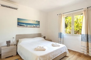 Ein Schlafzimmer mit einem Bett, einem Nachttisch, einer Wand-Klimaanlage, einem Fenster mit Vorhängen und einem Bild mit Strandmotiv.