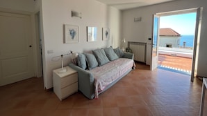 Una habitación con sofá, mesita de noche, lámpara y vista al mar por una puerta abierta.