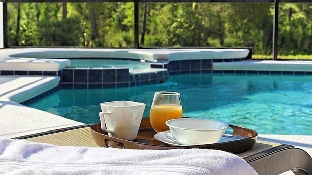 Un petit-déjeuner servi au bord de la piscine, avec un plateau contenant des tasses blanches, un verre de jus d’orange et un bol, donnant sur un jacuzzi et une piscine dont le bord est recouvert de carreaux.
