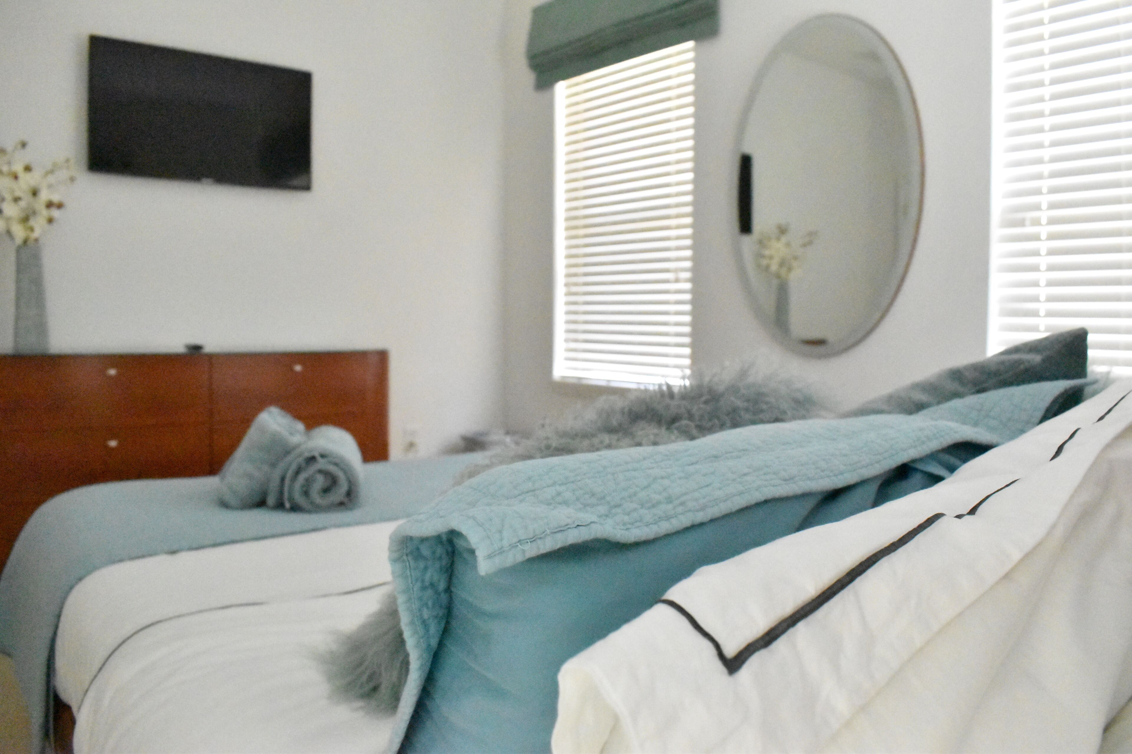 A neatly made bed with a blue blanket and pillows, a wooden headboard, a wall-mounted TV, and a window with blinds.
