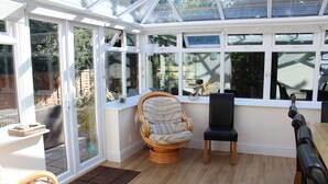 A conservatory with a glass door, a wicker chair, a black chair, and a wooden table. - New Forest cottage, sleeps up to 10. Looks directly on to the Forest  (Brockenhurst)