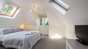 A bedroom with a sloped ceiling, skylight, a bed with a white comforter, a bedside table with a lamp, and a TV on a wooden dresser. - New Forest cottage, sleeps up to 10. Looks directly on to the Forest  (Brockenhurst)