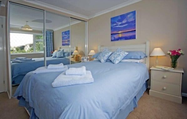 A bedroom with a large bed, a bedside table with a lamp, a mirror, and a painting on the wall. - 4-Star Family Home in Rock: private garden, walk to beach & pet friendly (Wadebridge)