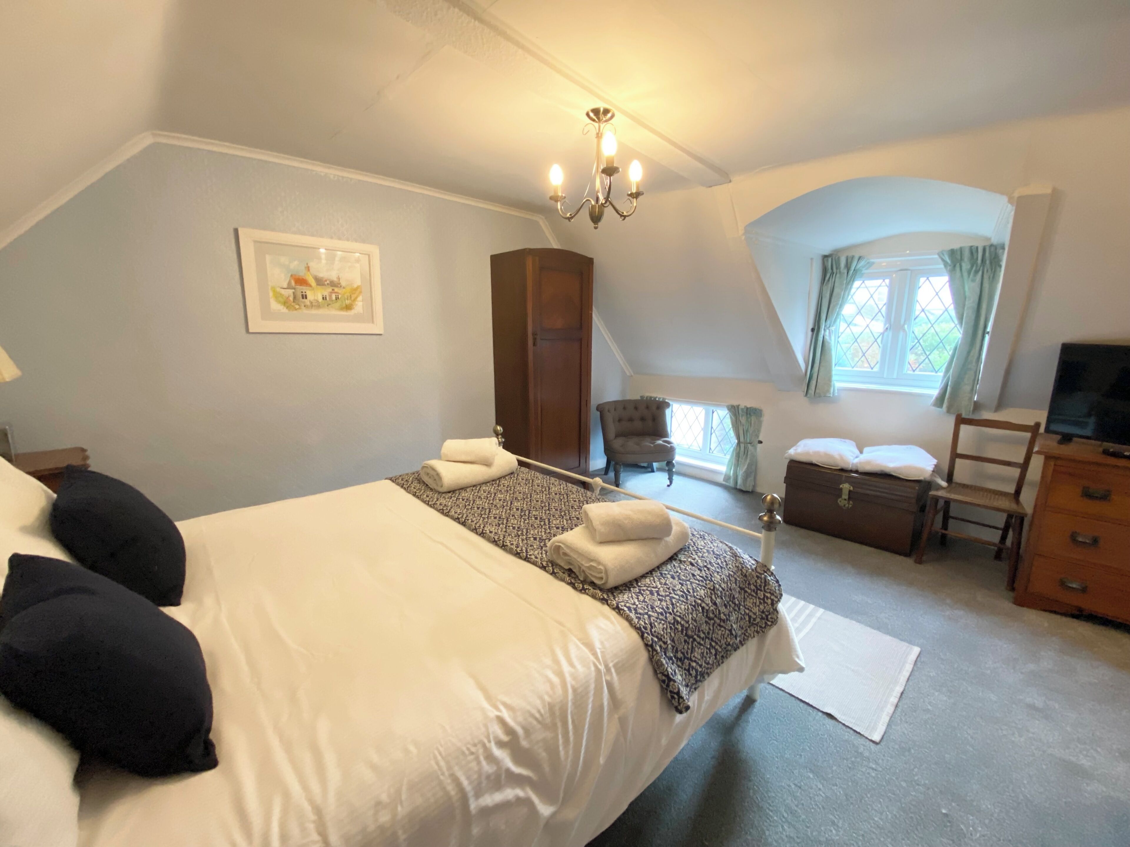 Une chambre à coucher avec un grand lit, une chaise, une commode, un téléviseur et une fenêtre avec des rideaux.
