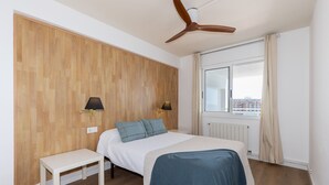 Un dormitorio con paneles de madera, una cama con manta azul, dos lámparas de noche, un ventilador de techo, una ventana con cortinas y vistas a edificios.