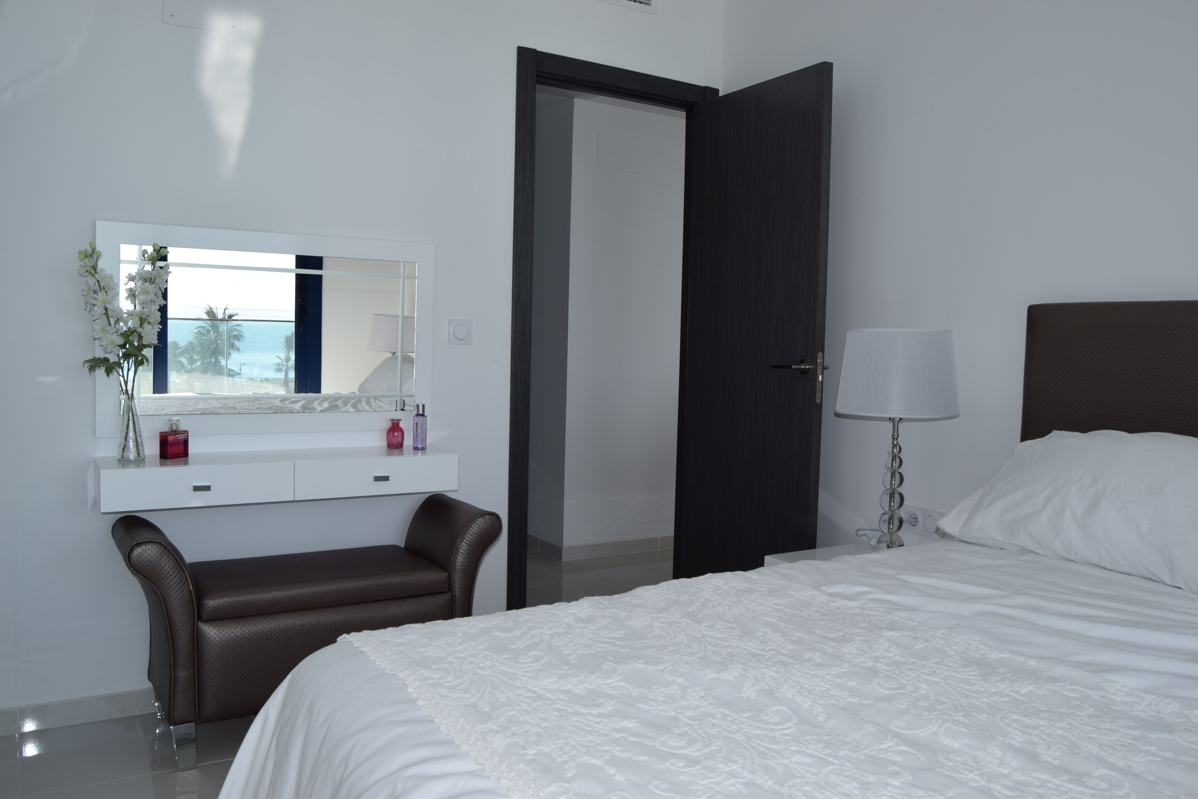 A hotel room with a bed, a bedside table with a lamp, a mirror, a bench, and a door.