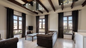 Ein modernes Wohnzimmer mit Sofa, Couchtisch und Fernseher. Große Fenster mit Vorhängen, Holzbalken an der Decke und ein Balkon, der durch die Fenster sichtbar ist.