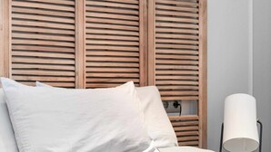 Un letto rifatto con lenzuola e cuscini bianchi, una testiera in legno con listelli orizzontali e una lampada da terra a destra.
