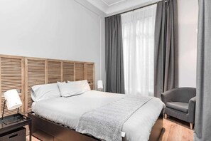 Une chambre d’hôtel moderne avec une tête de lit en bois, un lit simple, un fauteuil gris et une table de chevet avec une lampe.