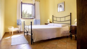 Un dormitorio con cama, mesitas de noche, una silla, ventana con cortinas y un cuadro en la pared.