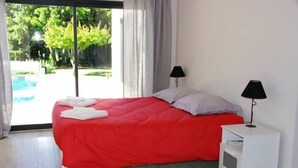 Une chambre à coucher avec un lit, deux lampes de chevet, une table de chevet blanche et une vue sur une piscine et la verdure environnante.