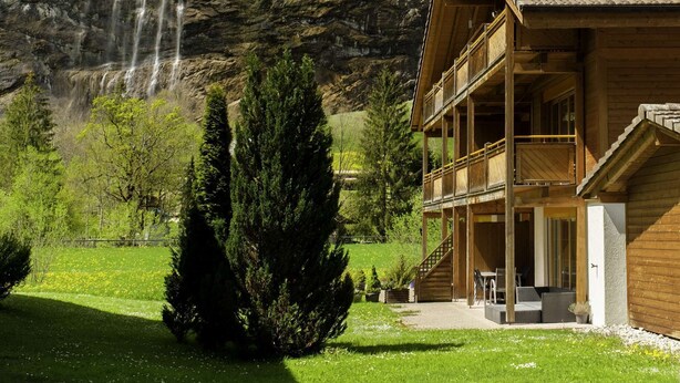 A wooden lodge with a balcony overlooking a waterfall and forested mountains.