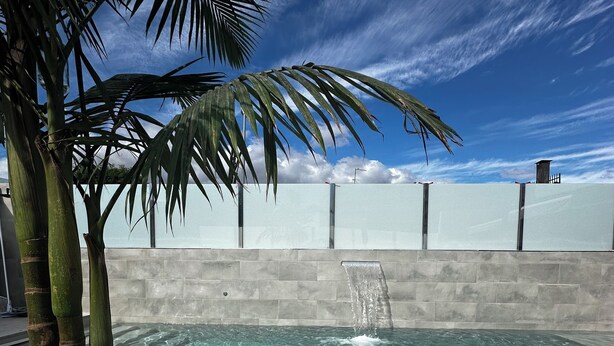 A swimming pool with a water feature, surrounded by a concrete deck and palm trees.