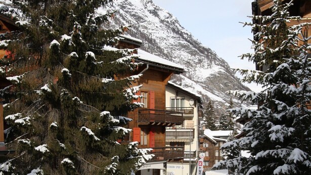 A snow-covered street with buildings, including a multi-story structure with balconies. - High Standard Renovated Apartment for up to 8 persons, in the Centre (Zermatt)