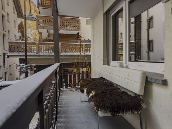 A narrow balcony with a fur-covered bench, a railing, and a view of a building with balconies and string lights.