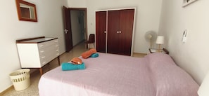 Een slaapkamer met een bed, een dressoir, een kast, een stoel, een ventilator en een lamp.