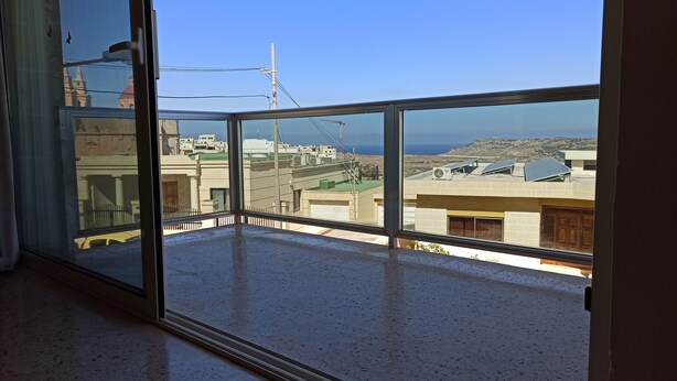 A room with a large window overlooking a cityscape with buildings and a distant view of the sea. - Apartment with Sea View (Flat 5) (Triq L-Gherien, Mellieha)
