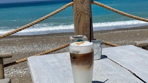 A glass of iced coffee on a wooden table by the beach with a rope barrier and a wooden post in the background.