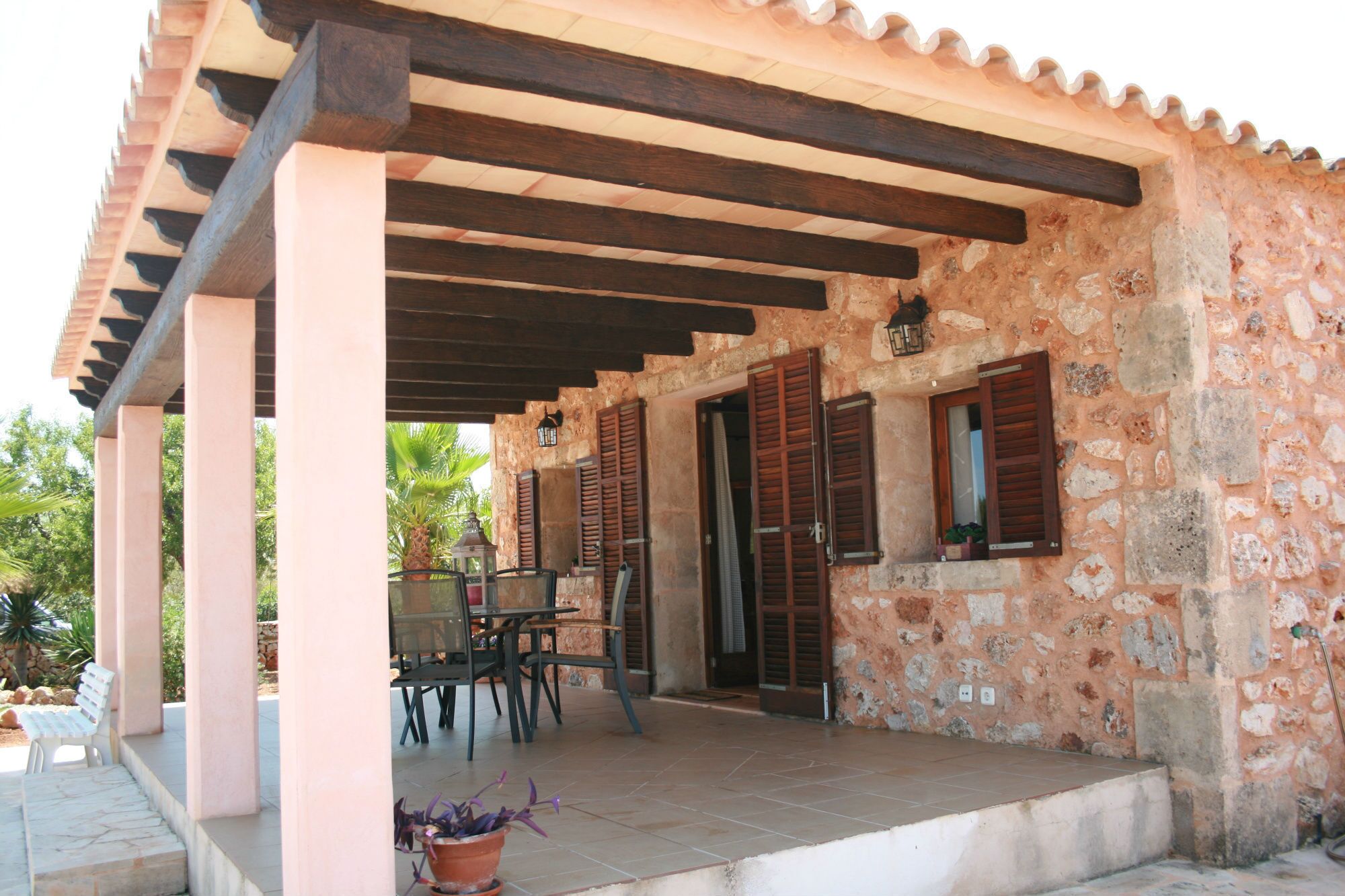 A covered outdoor seating area with stone walls and wooden beams, featuring a table and chairs.