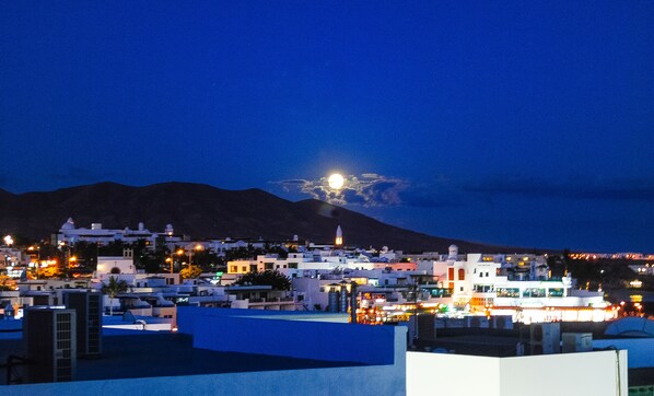 Une vue nocturne d’une ville, avec une montagne imposante en arrière-plan et une lune brillante dans le ciel.