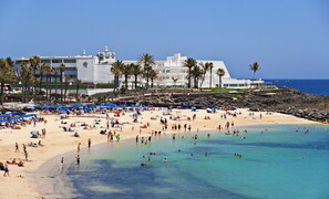 Une plage avec une eau turquoise et limpide, des palmiers et, au loin, un grand bâtiment doté de nombreux balcons.