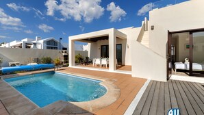 Un espace piscine extérieur moderne, doté d’une terrasse en bois, d’un coin salon abrité et d’un ciel d’un bleu limpide.