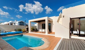 Un espace piscine extérieur moderne, doté d’une terrasse en bois, d’un coin salon abrité et d’un ciel d’un bleu limpide.