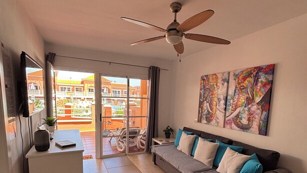 Una sala moderna con sofá, mesa de centro, televisor y ventilador de techo. Hay un balcón con vista a edificios.